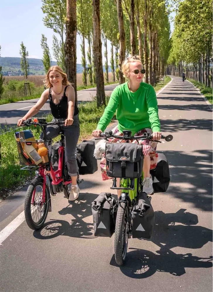 Zwei Radfahrerinnen auf E-Bikes mit Packtaschen auf einem Radweg in einer Pappelallee