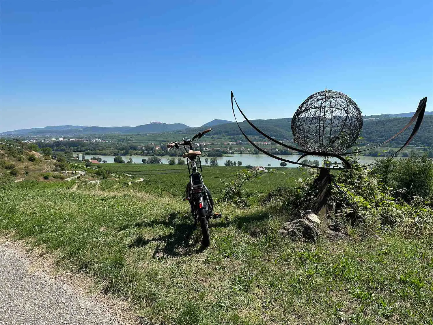 E-Bike auf einer Wiese neben einer Kugel-Skulptur aus Metall, dahinter Weingärten und die Donau