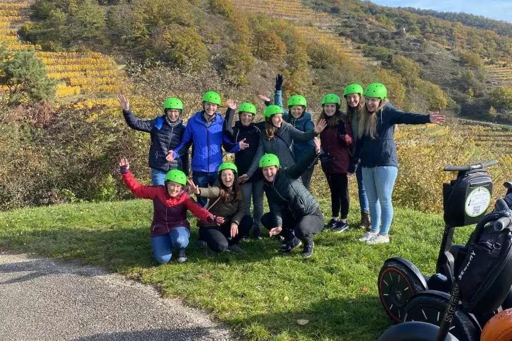 Gruppe mit grünen Helmen jubelt auf einer Wiese vor herbstlichen Weinbergen, daneben Segways