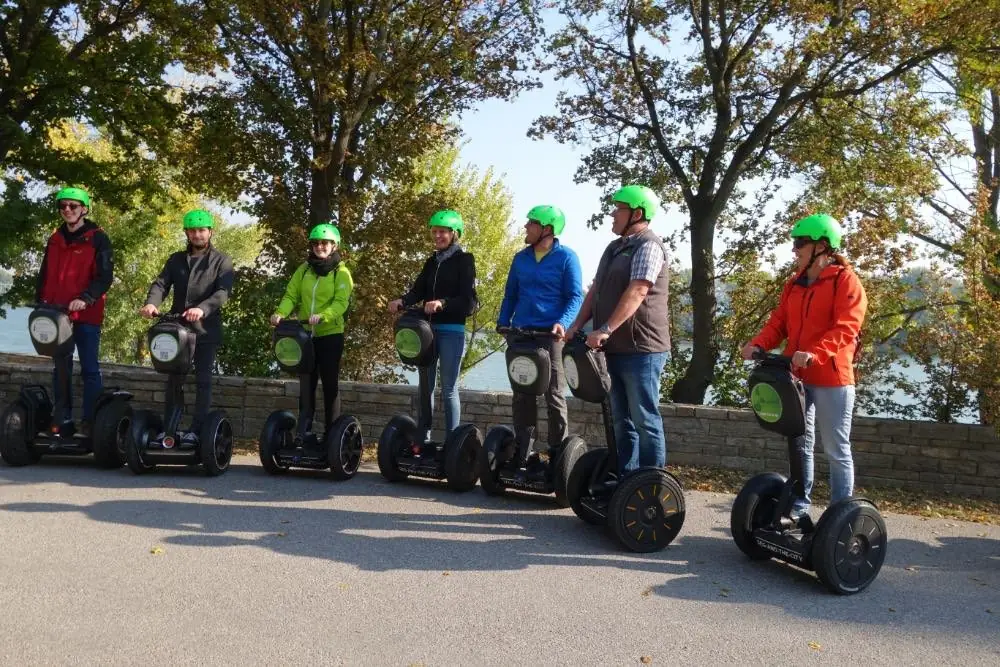 Sieben Segway-Fahrer mit grünen Helmen nebeneinander an einer Mauer unter Bäumen